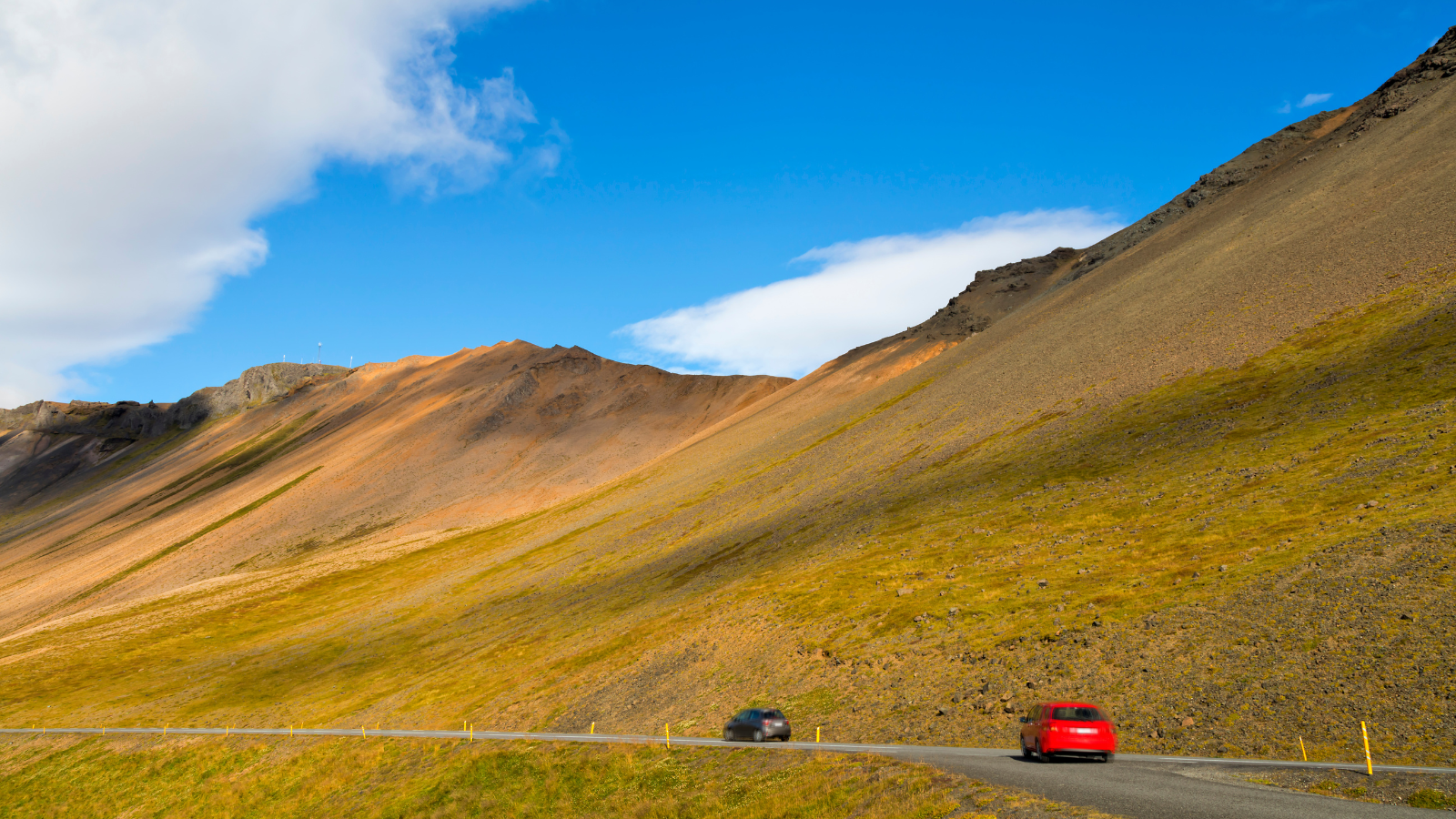 Blue skies in Iceland over two cards driving on a road beside a grassy peak.