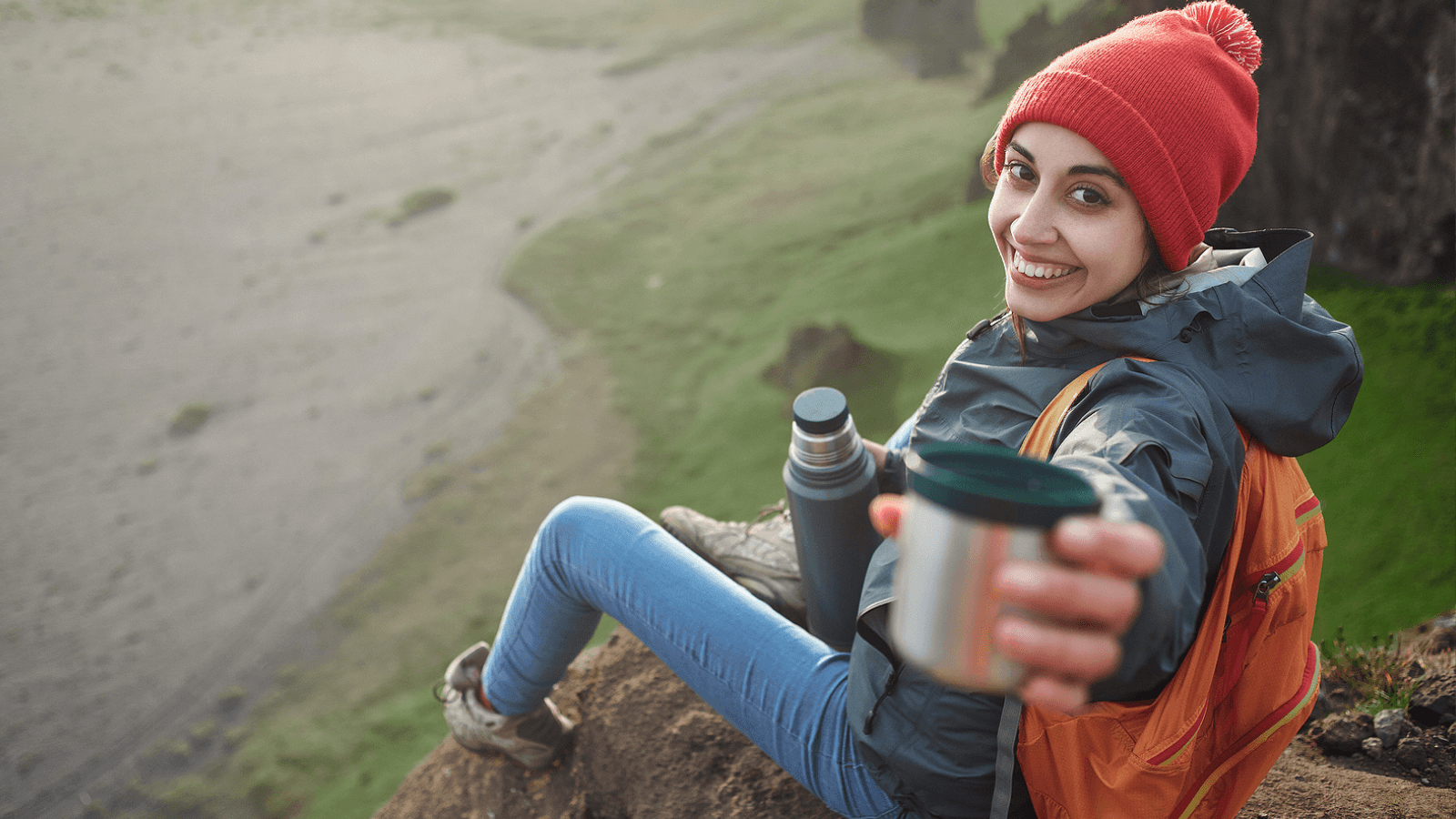 Woman sitting on a cliff in Iceland holding a coffee thermos cup towards the camera.